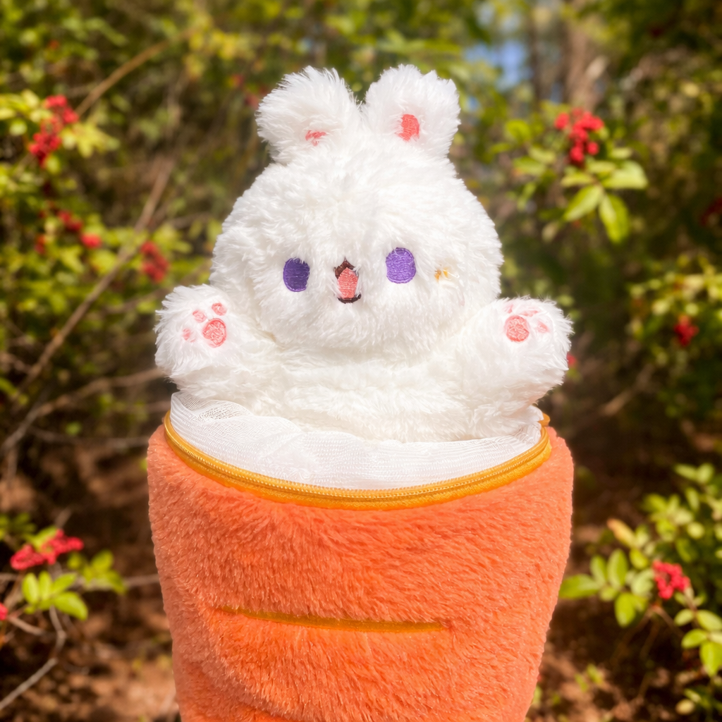 Plush toy resembling a white creature with bunny ears in an orange carrot-shaped container against a natural background.